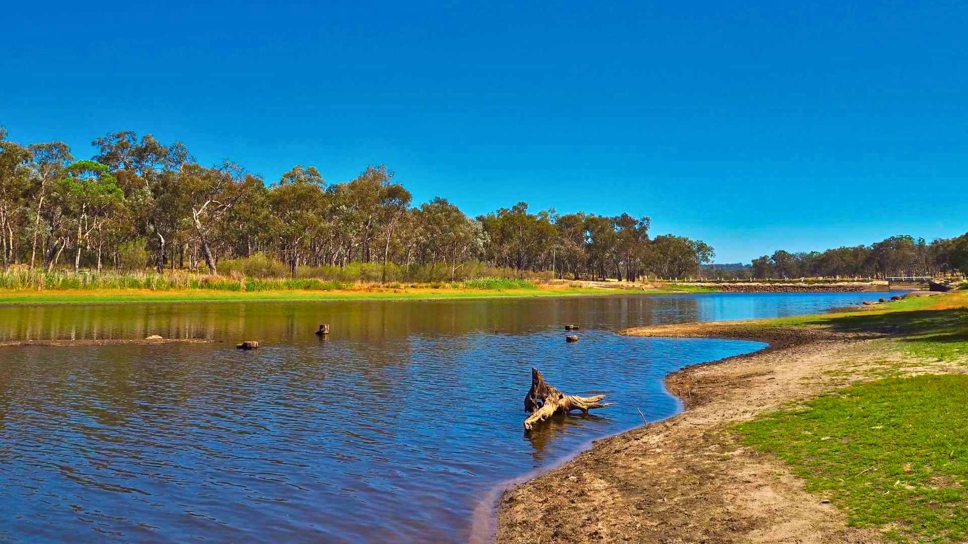 Narrow reservoir with sandy banks, tree line and bright blue sky.