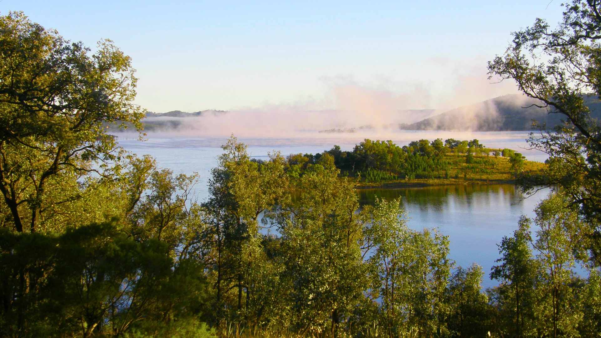 Morning mist rising over Glenlyon Dam with tree-lined shores and distant hills.