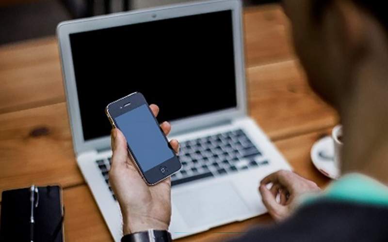 photo of open laptop on a table and the arm of a person holding a mobile phone