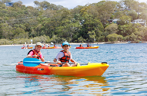 Children sitting in a colored canoe on a lake, with other manned canoes in the background and trees surrounding the lake