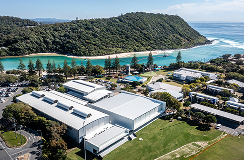 Aerial view of the venues buildings, with a grassed foreground and the river mouth and green hills in the background.